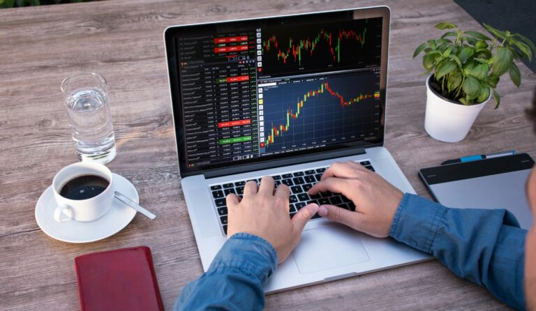 Handen typen op een laptop met beurskoersen en candlestick-grafieken, met een kop koffie, een glas water en een plantje op een houten tafel